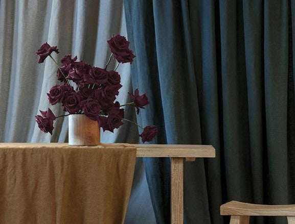 A bunch of roses in a vase on top of a wooden shelf with linen curtins in the background