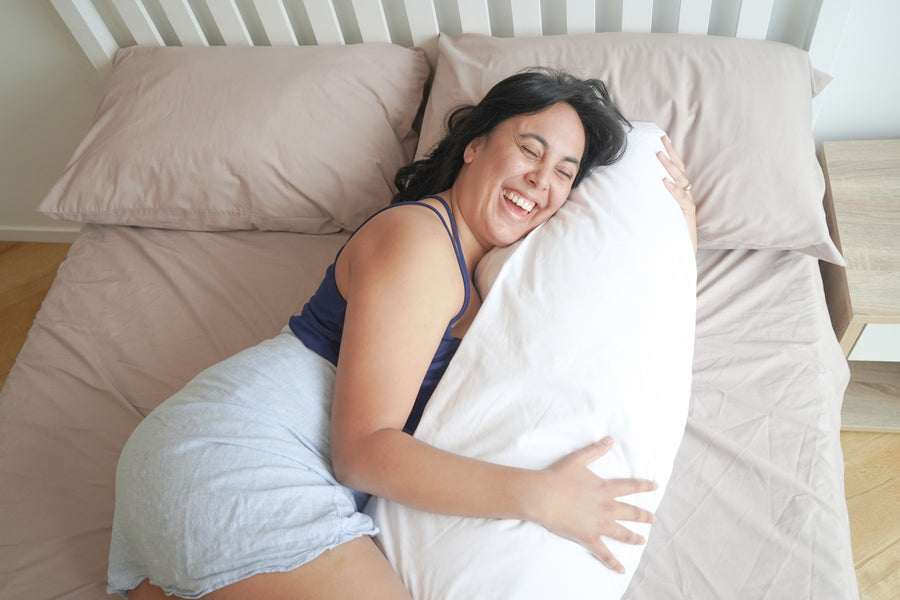 Woman laughing whilst lying on a bed cuddling a Kind Face Cloud Wool Body Pillow 